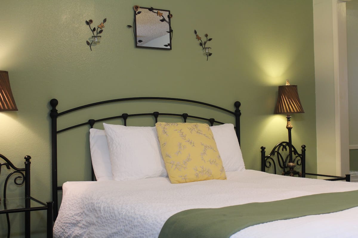 A neatly made bed with white linens and a yellow pillow, a black metal headboard, and two wall-mounted lamps on either side.