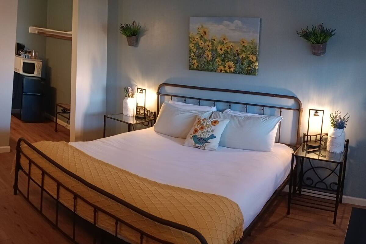 A bedroom with a bed, bedside tables, a nightstand, a wooden bench, a small table with a jar, and a wall-mounted plant.