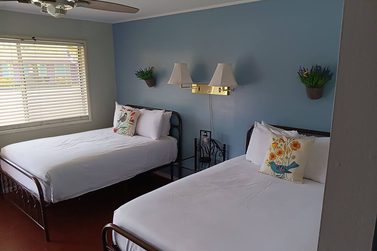 A room with two beds, a ceiling fan, a window with blinds, and wall-mounted lamps.