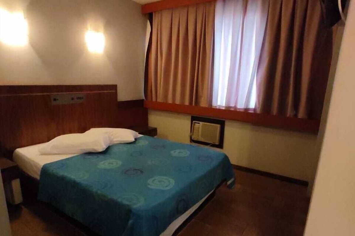 A hotel room with a bed, wooden headboard, a window with curtains, and a wall-mounted TV.