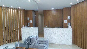 Zona de recepción con mostradores de mármol, un separador de madera y un área de estar con sofás grises.