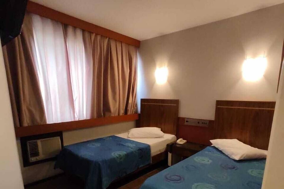 A hotel room with two single beds, a window with curtains, a wall-mounted heater, and a nightstand with a phone.