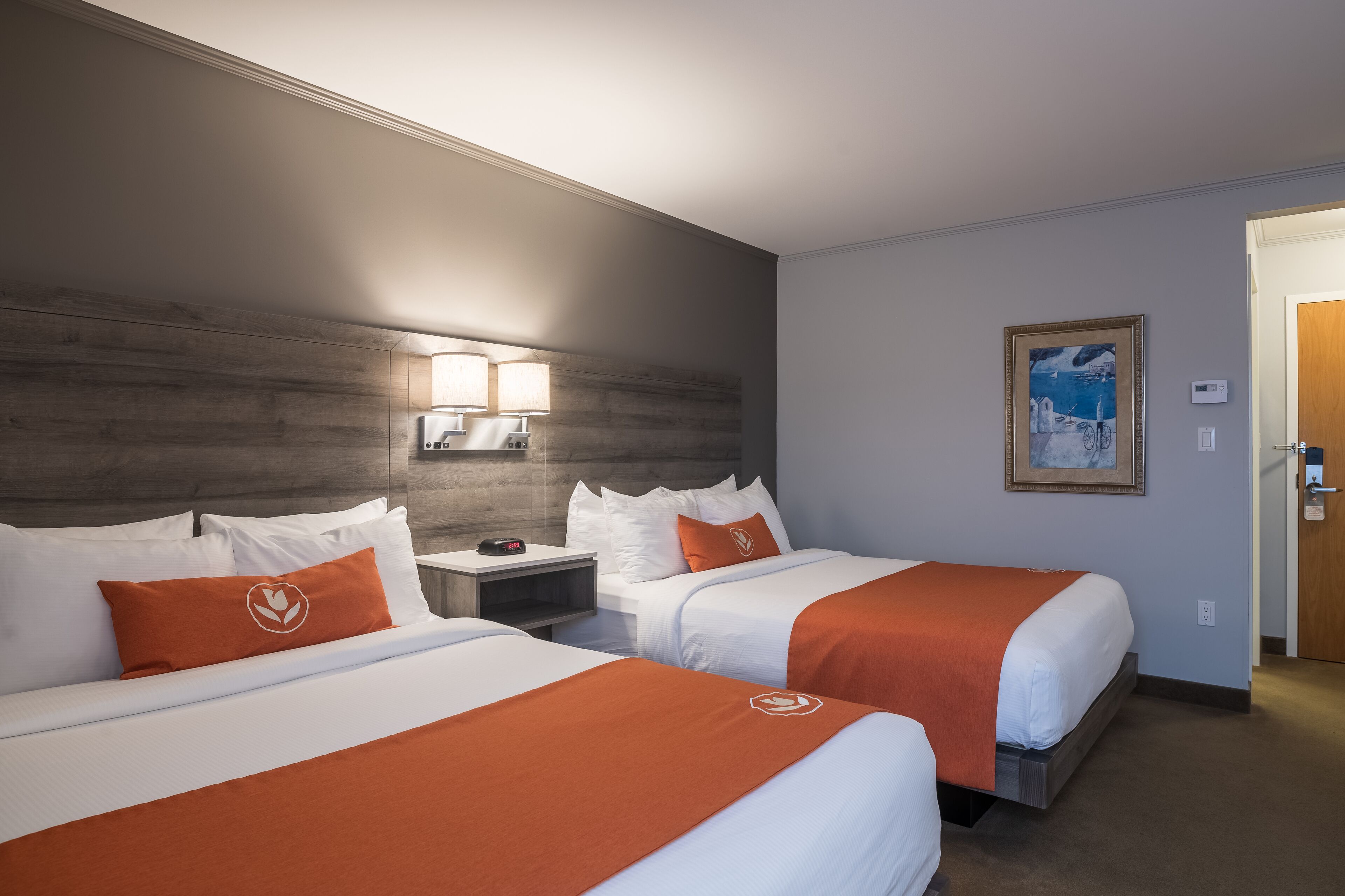 Habitación de hotel con dos camas, cada una con ropa blanca y detalles naranja, una mesita de noche con lámpara, un cuadro en la pared y una puerta a la derecha.