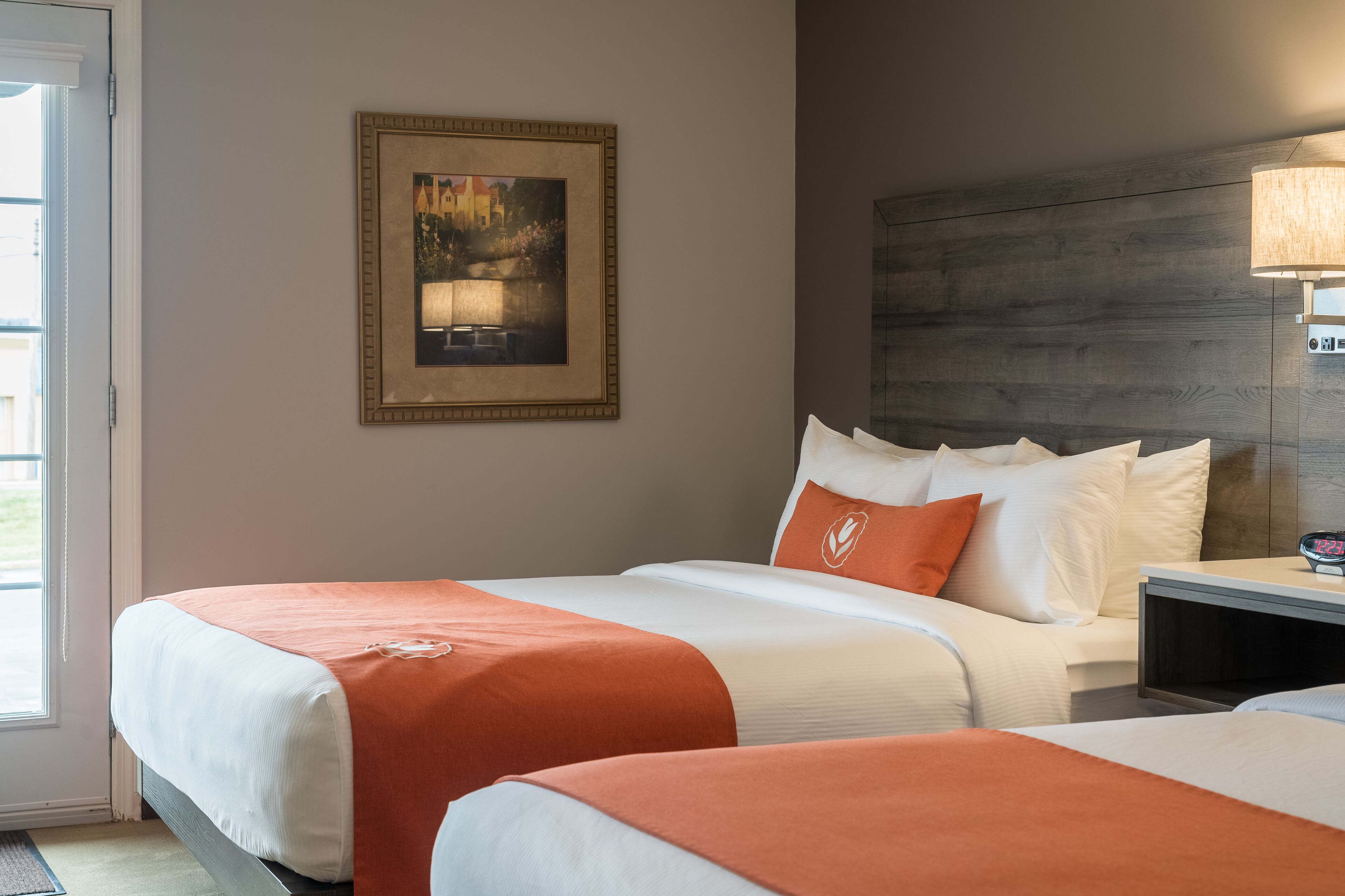 A hotel room with two beds, a framed painting on the wall, a nightstand with a lamp, and a view of the outdoors through a door.