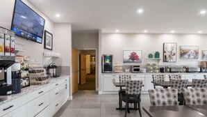 Una cocina moderna con armarios blancos, un comedor con sillas estampadas y un televisor en la pared mostrando un pronóstico del tiempo.