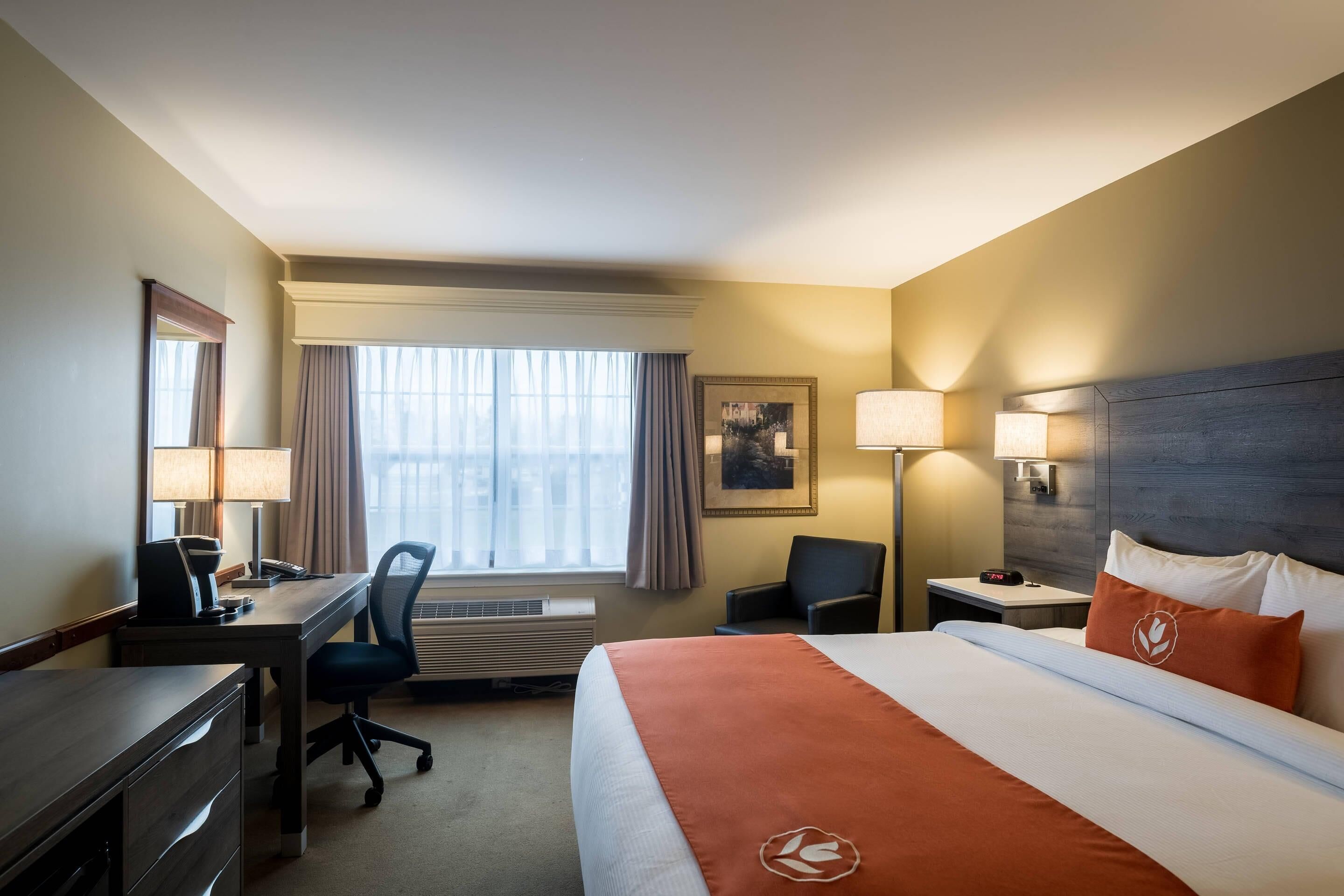 A hotel room with a bed, desk, chair, lamp, and a window with curtains.