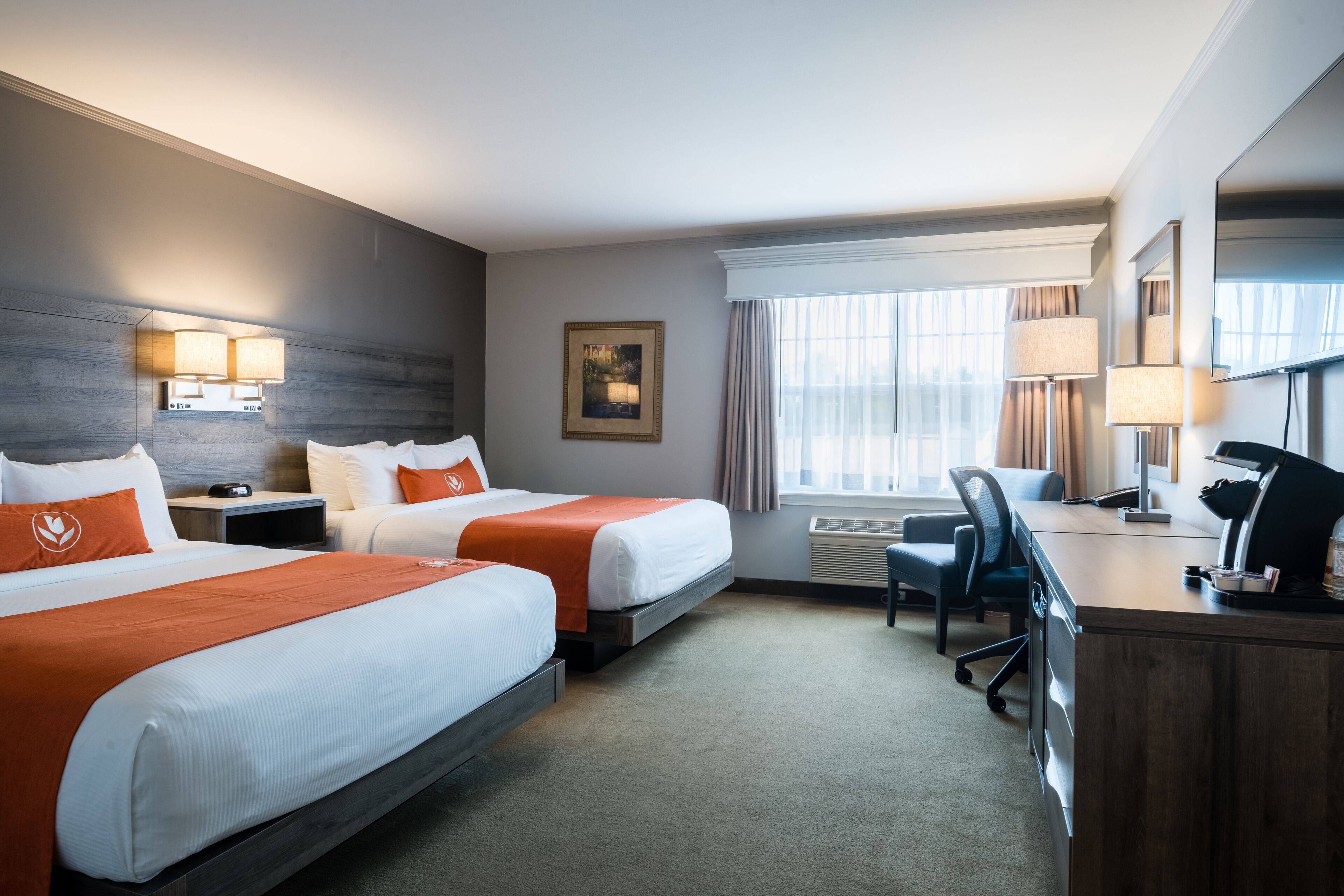 A hotel room with two beds, a desk, a chair, a desk lamp, a picture on the wall, and a window with curtains.