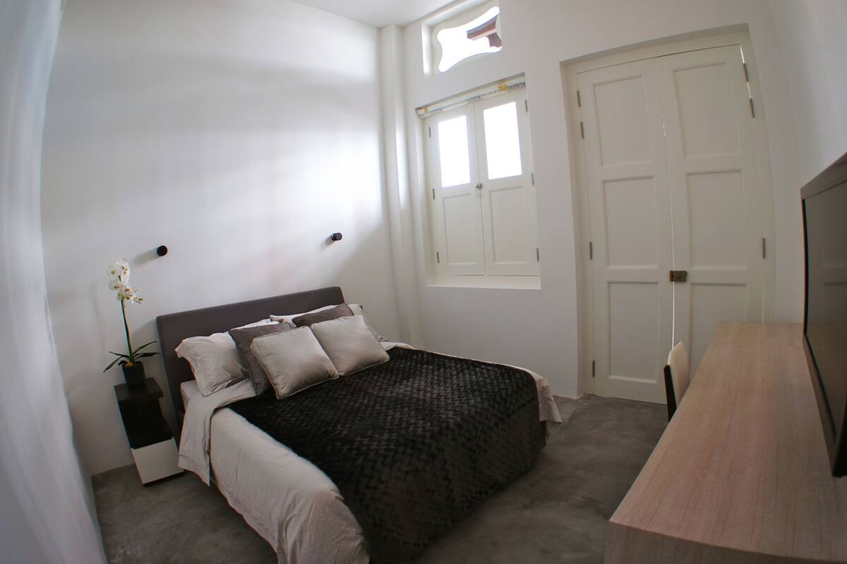 A bedroom with a bed, bedside table, TV, and a window.