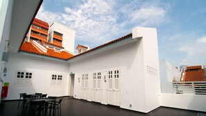 Un pasillo de hotel con una larga fila de taquillas blancas, un comedor con mesas y sillas, y vistas a edificios vecinos con tejados rojos.