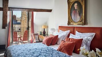 A hotel room with a bed, a painting, a balcony with red chairs, and a view of buildings.