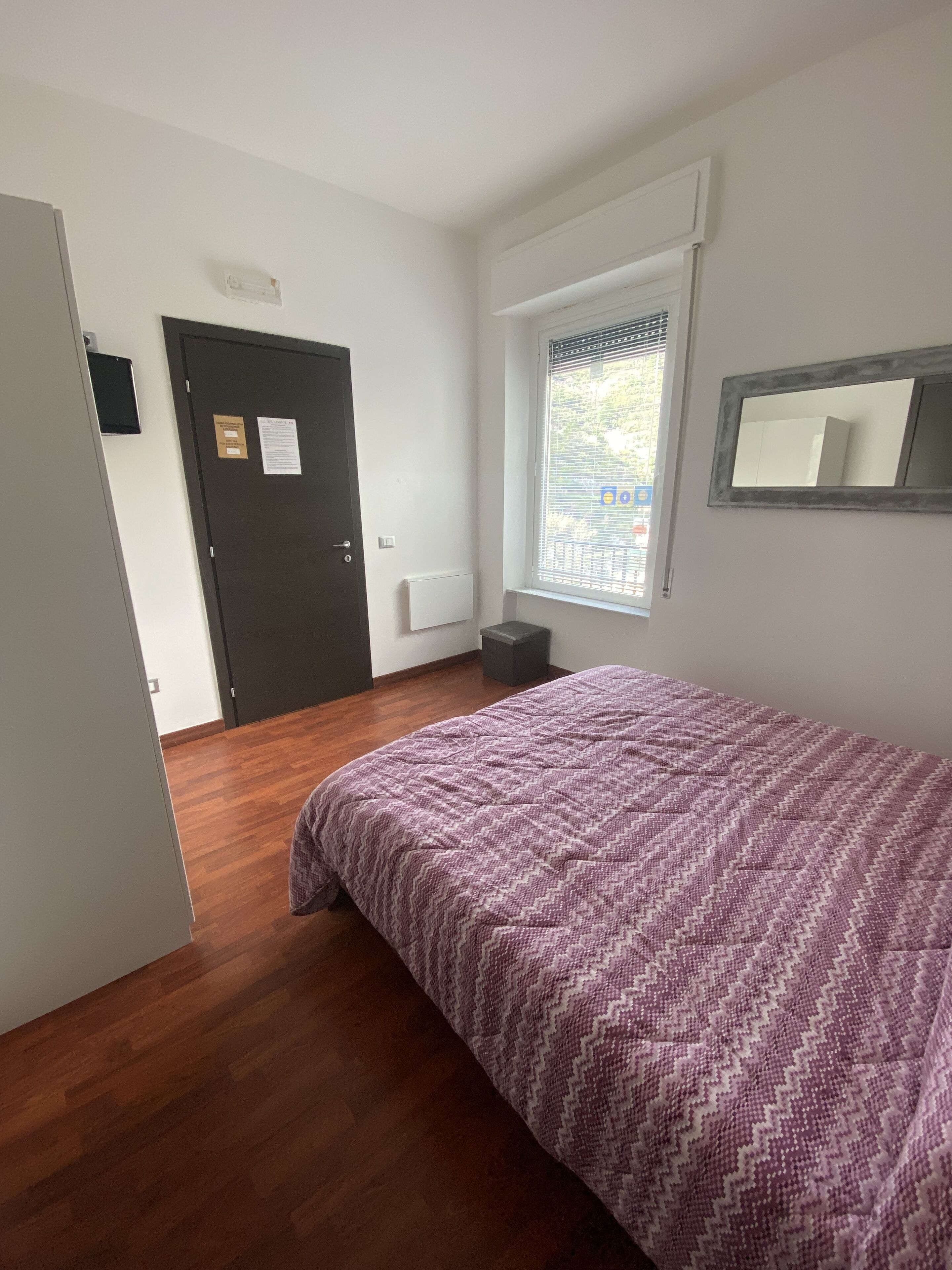 A bedroom with a wooden floor, a bed with a patterned bedspread, a window with blinds, a door with a notice, and a small table.