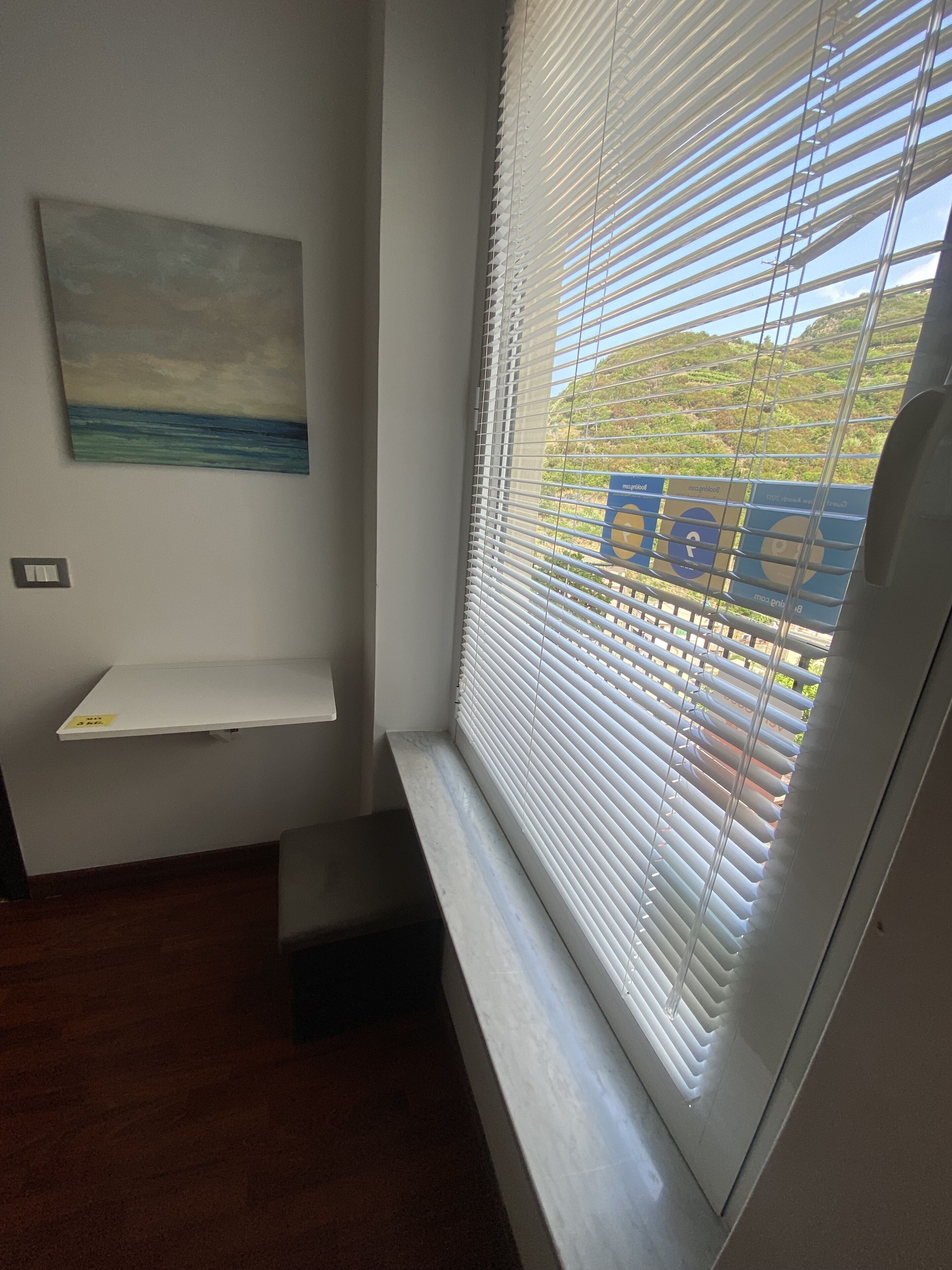 A room with a window with blinds, a small shelf, and a painting on the wall.