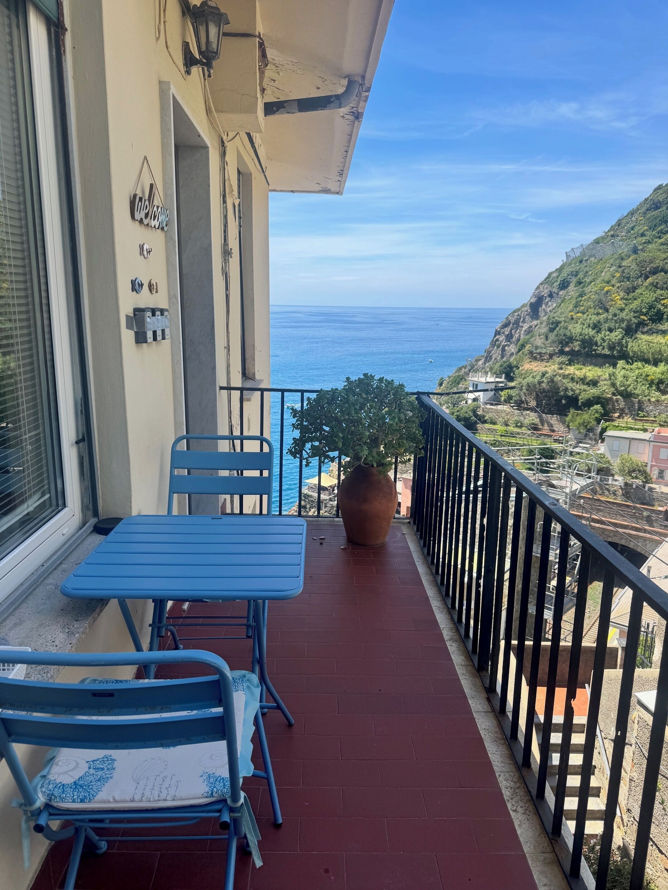 A balcony with a table and chairs, a potted plant, and a view of the sea and hills.