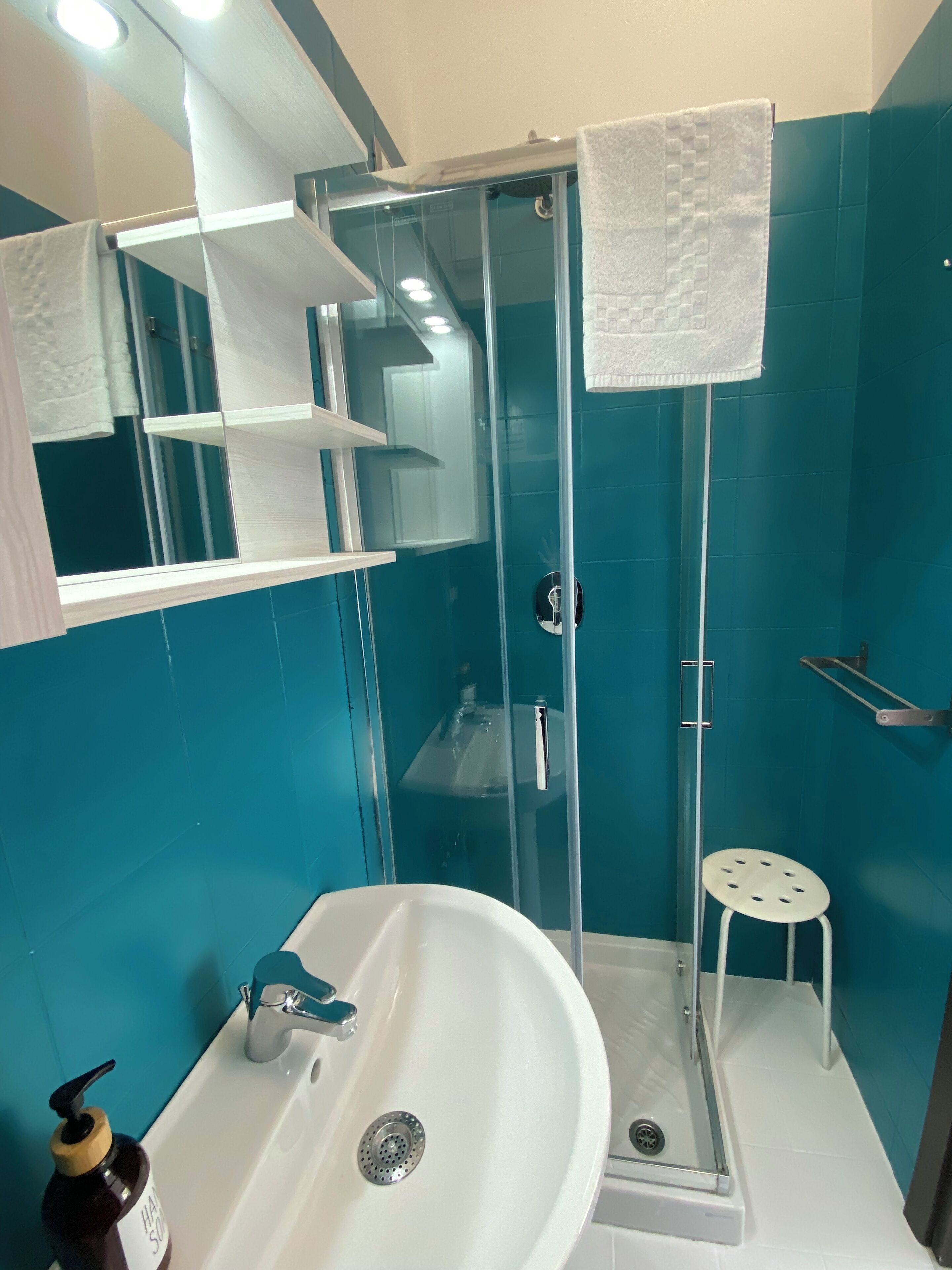 Ein Badezimmer mit einem weißen Waschbecken, einer Duschkabine, einem weißen Hocker und einem Spiegel mit Ablageflächen.