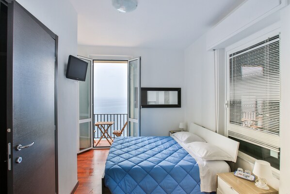 A bedroom with a bed, a TV, a window with blinds, and a balcony.