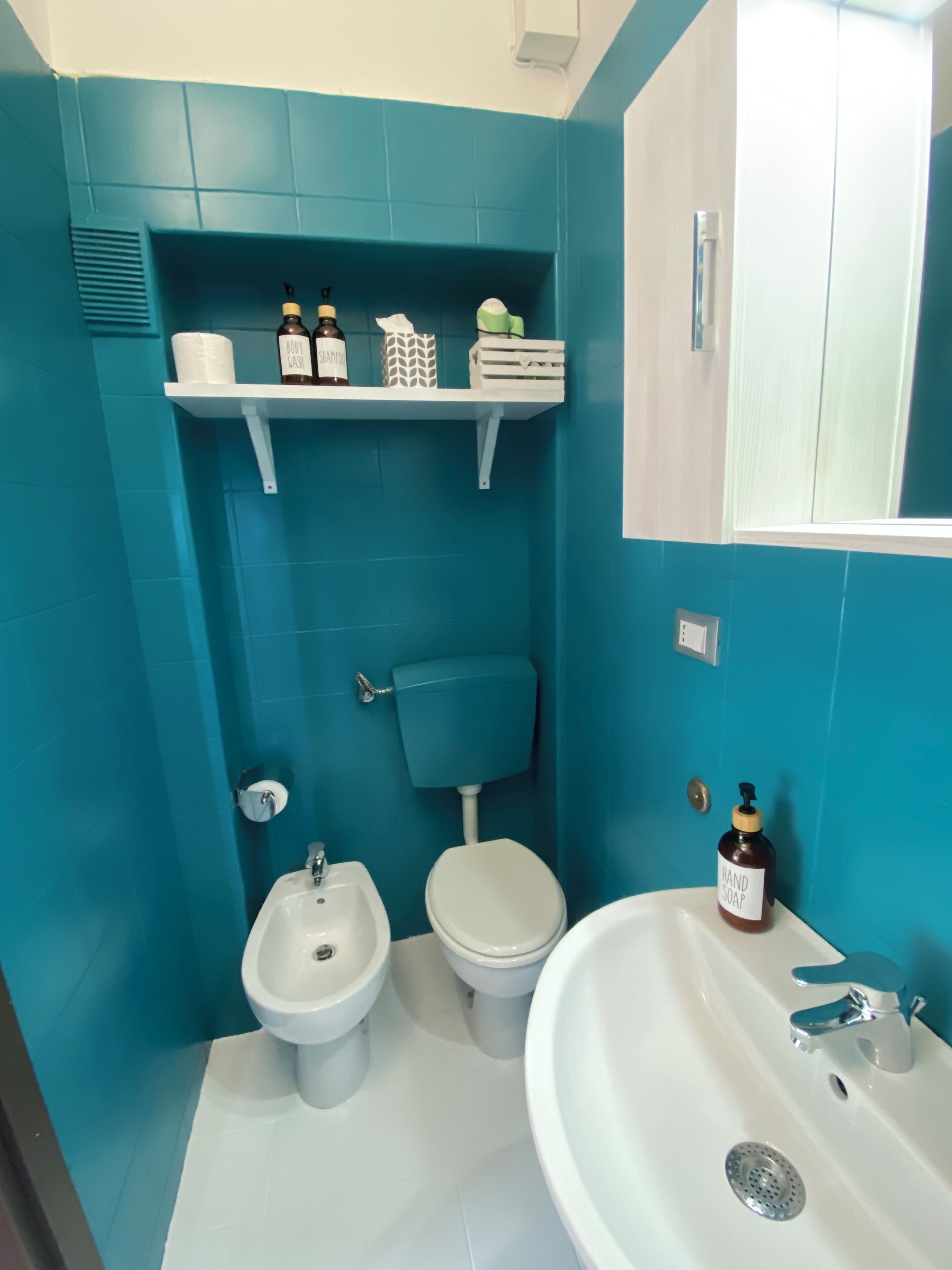 A bathroom with a toilet, bidet, and sink, with teal walls and white fixtures.
