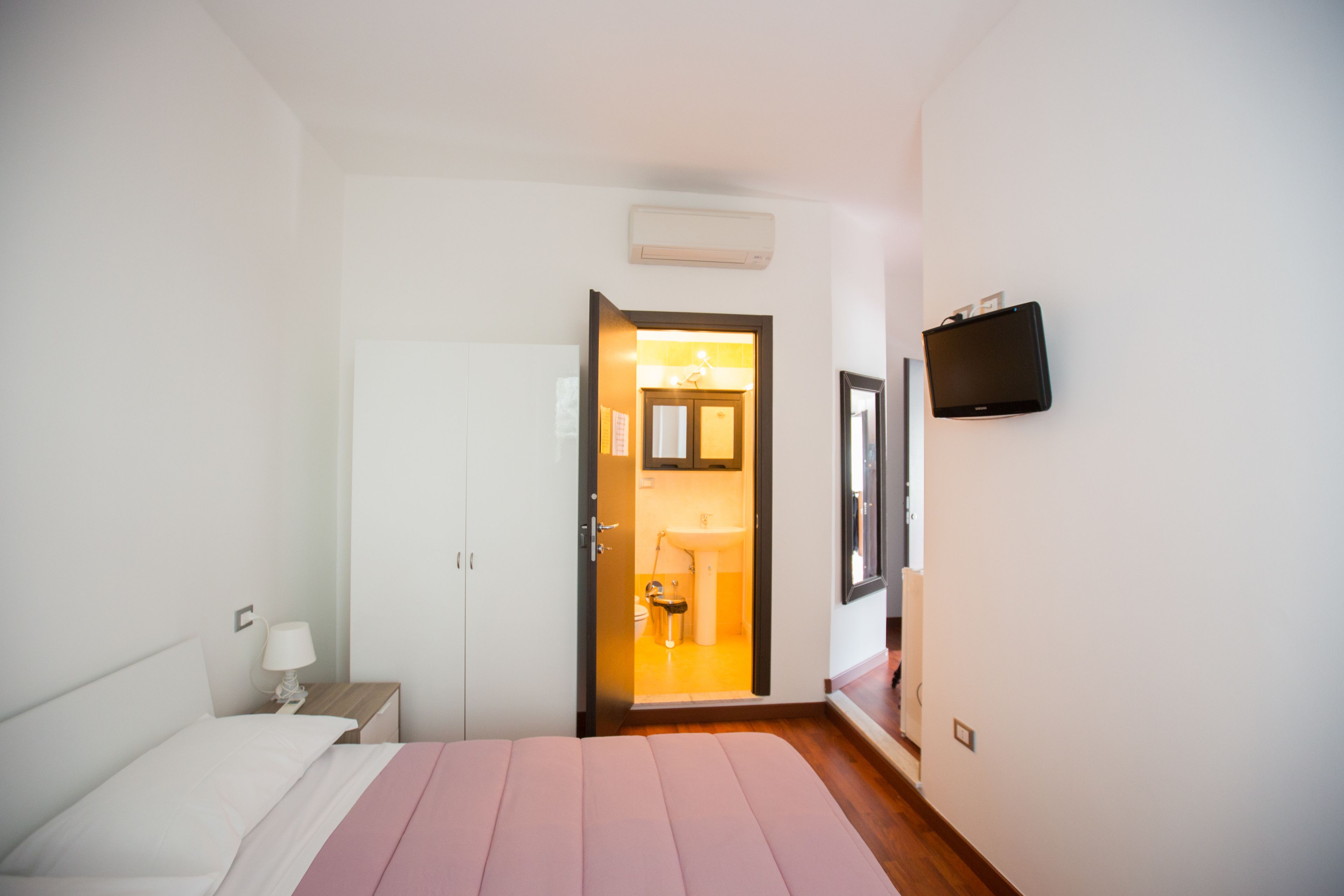 Un dormitorio con cama, mesita de noche, televisor montado en la pared y puerta abierta que da al baño.