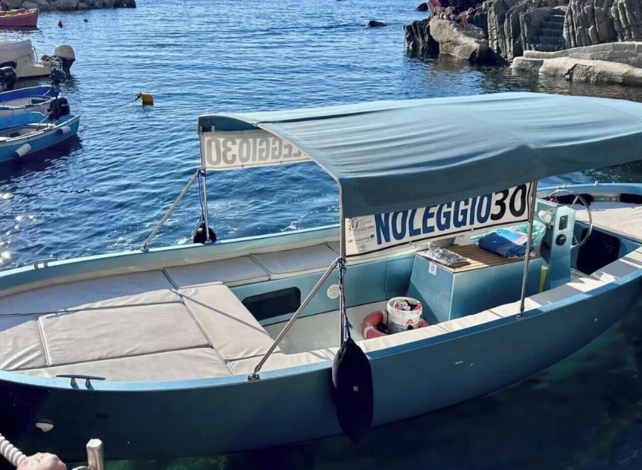 A small boat with a canopy and the number "NOLEGGIO30" on it, docked at a marina with other boats and rocky structures in the background.
