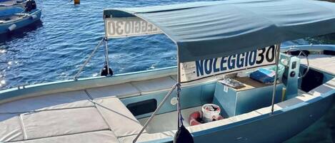 A small boat with a canopy and the number "NOLEGGIO30" on it, docked at a marina with other boats and rocky structures in the background.