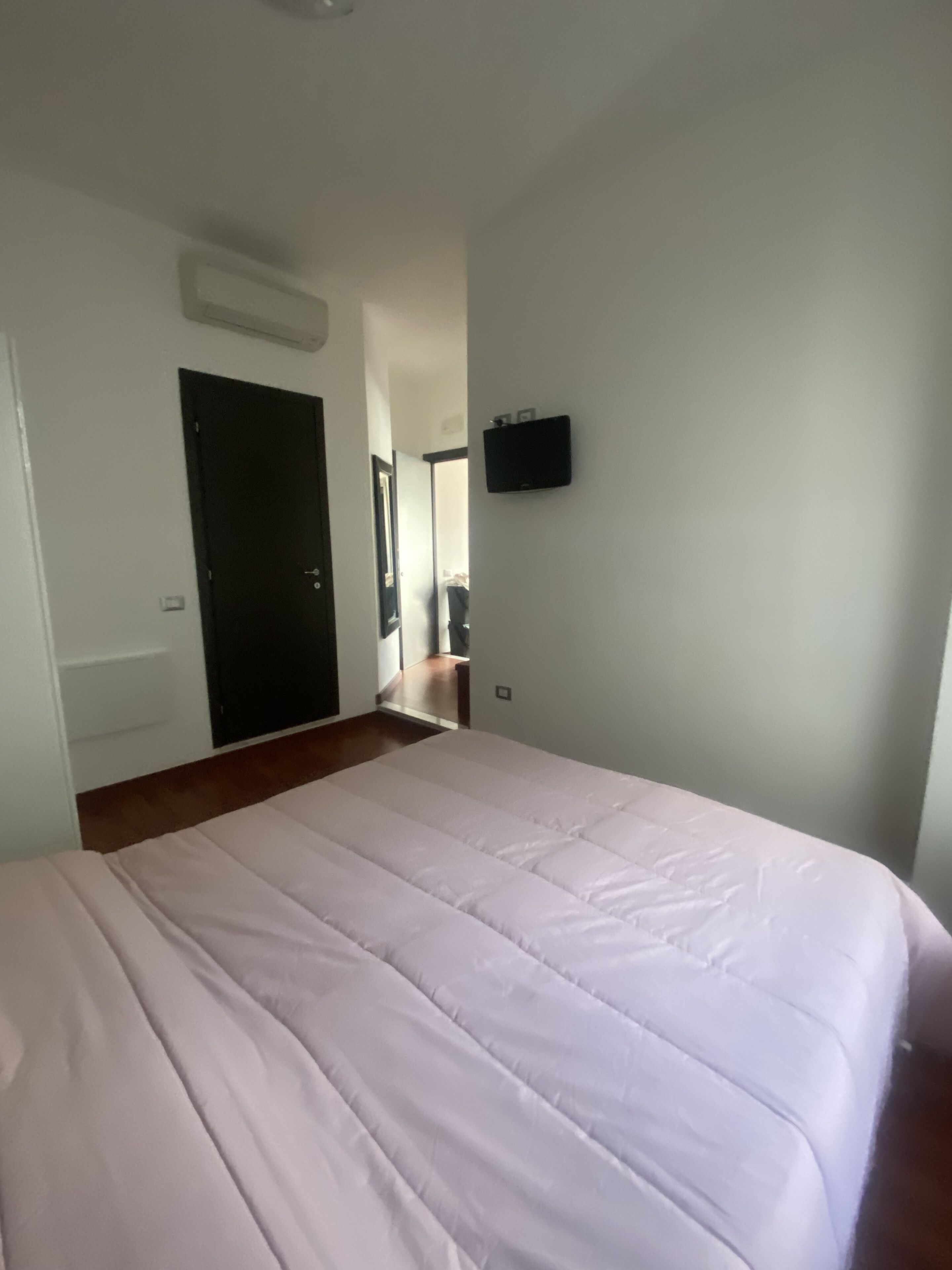 A bedroom with a bed, a door, a wall-mounted TV, and a hallway leading to another room.