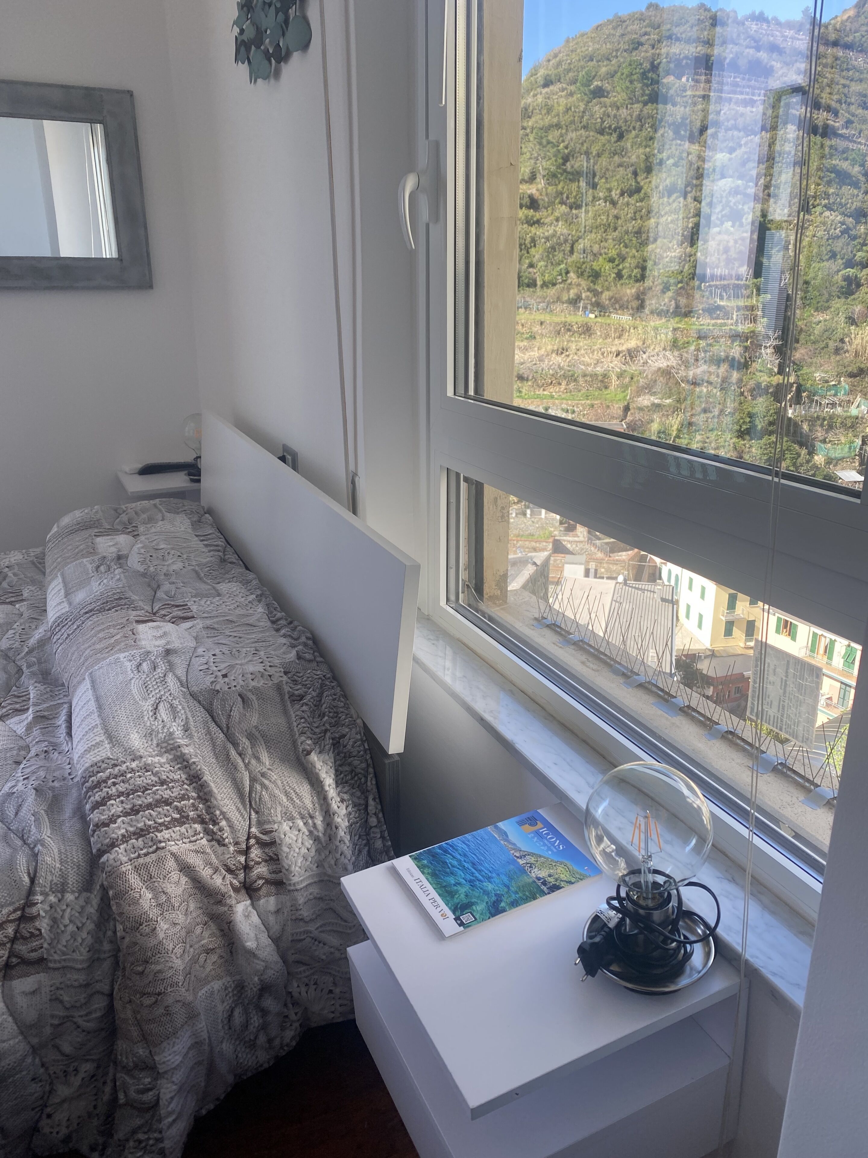 A bedroom with a bed, a bedside table with a book and a lamp, and a window with a view of a hillside.
