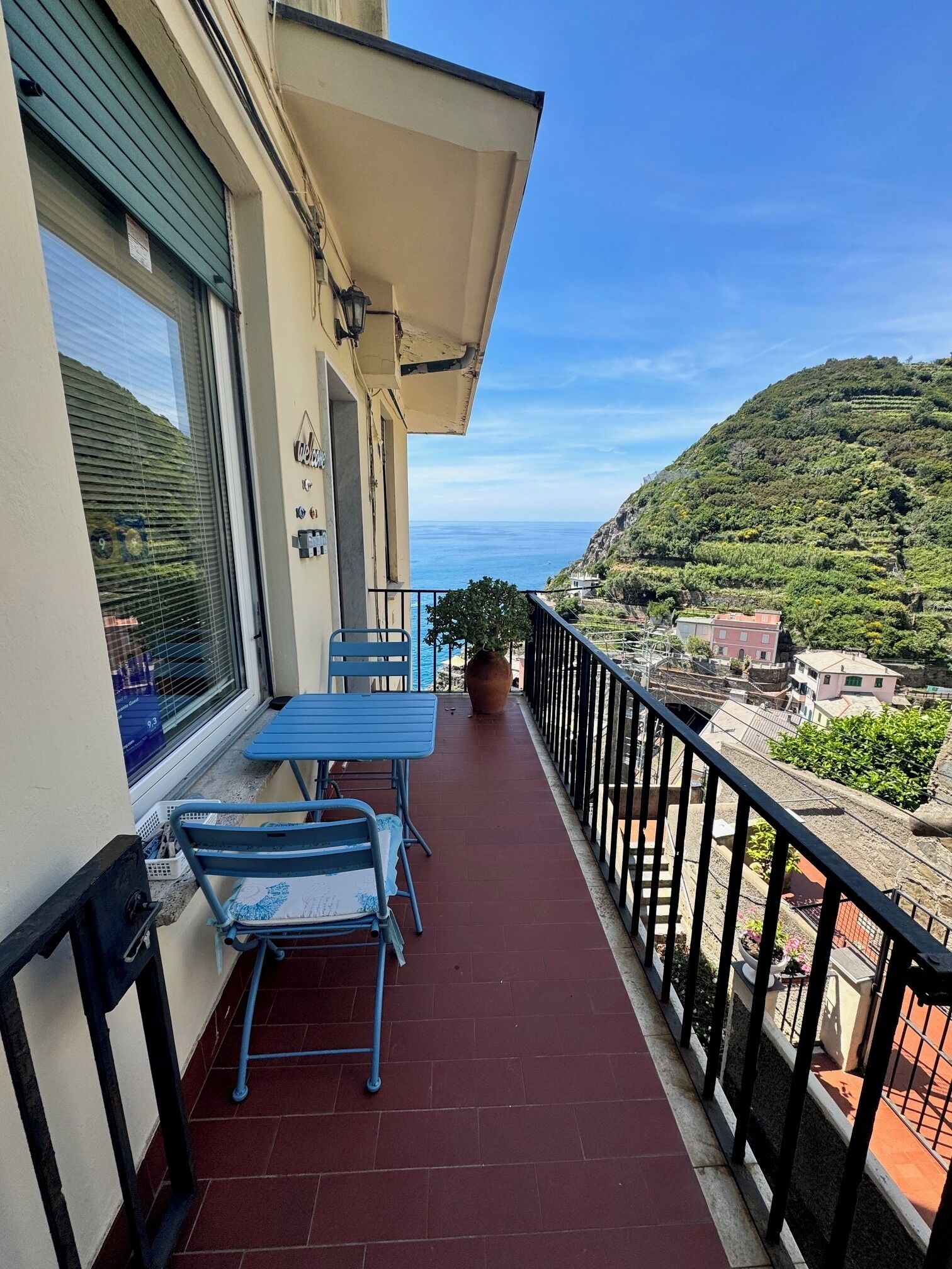 A balcony with a table and chairs, a potted plant, and a view of the sea and hills.
