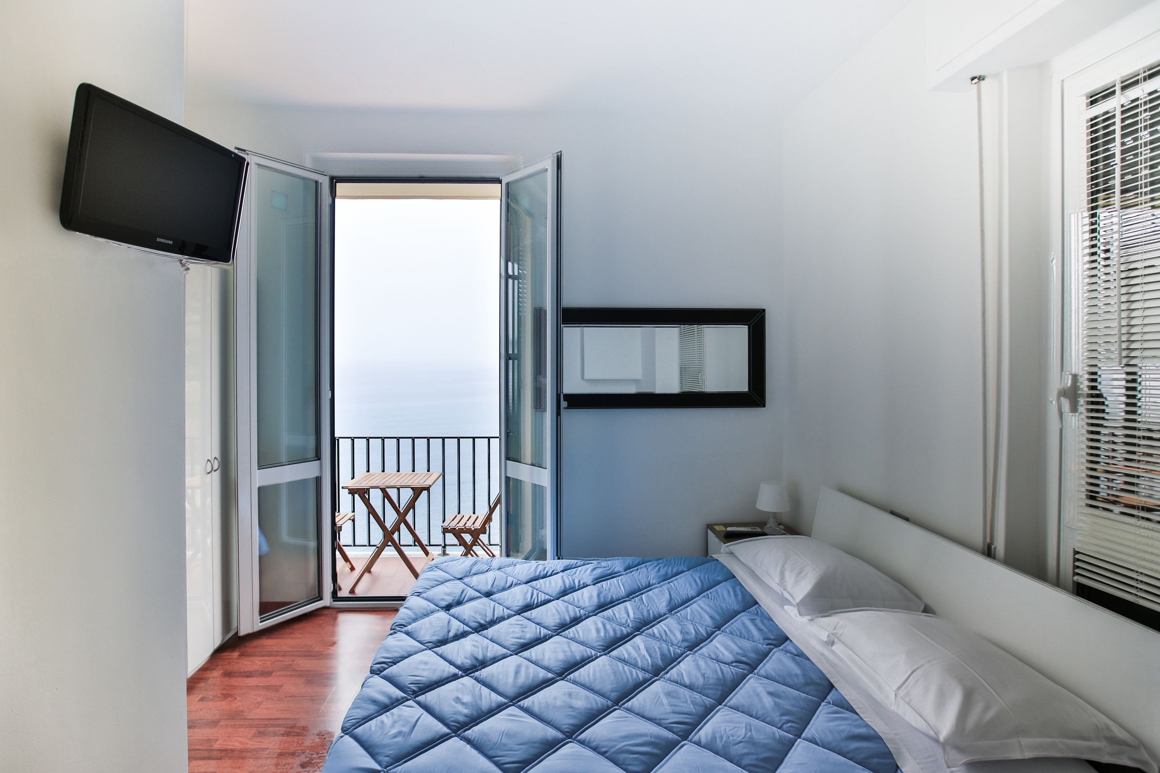 Un dormitorio con cama, televisión montada en la pared, ventana con persianas y balcón con sillas.