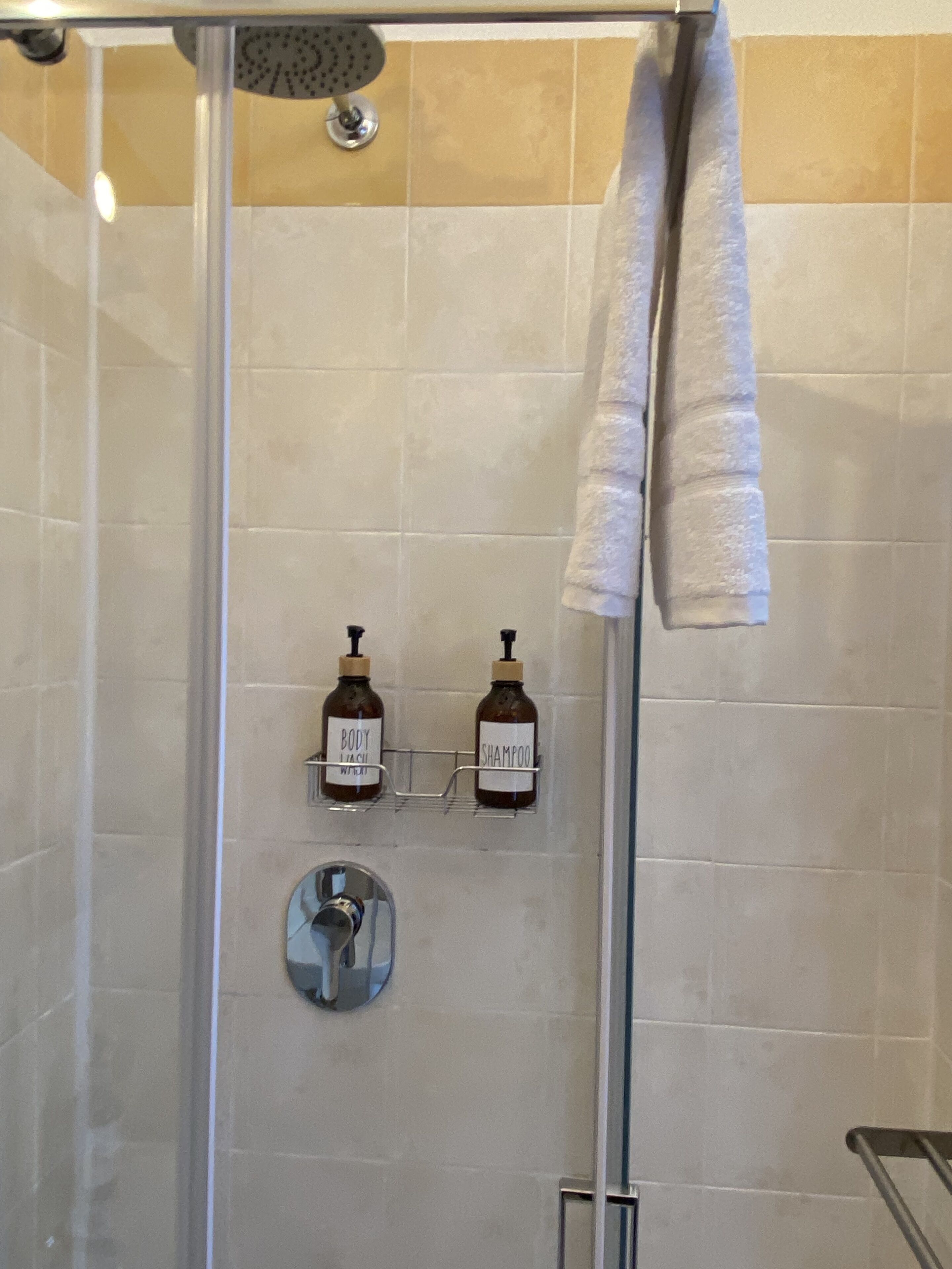 Ducha envuelta en vidrio con cabezal de ducha manual, dos botellas de jabón corporal y una toalla colgada en la pared.