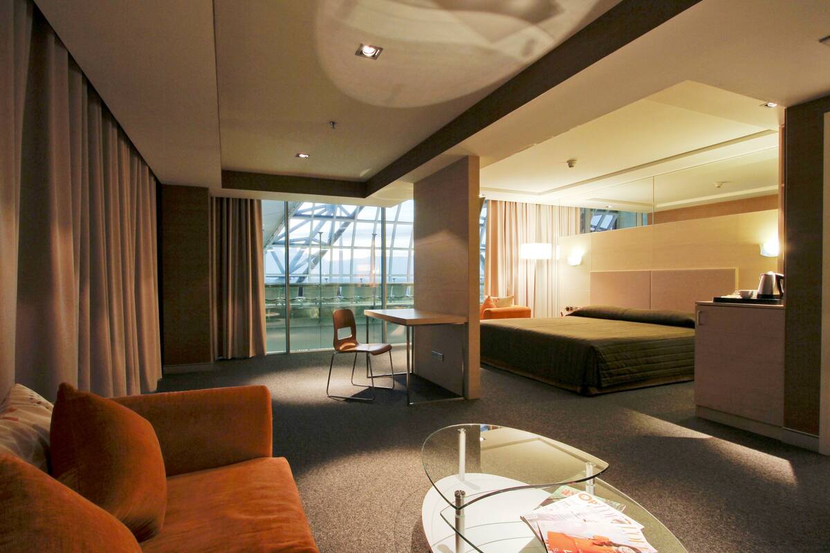 A modern hotel room with a glass partition, a sofa, a small table, a bed, a desk, and a kitchenette.