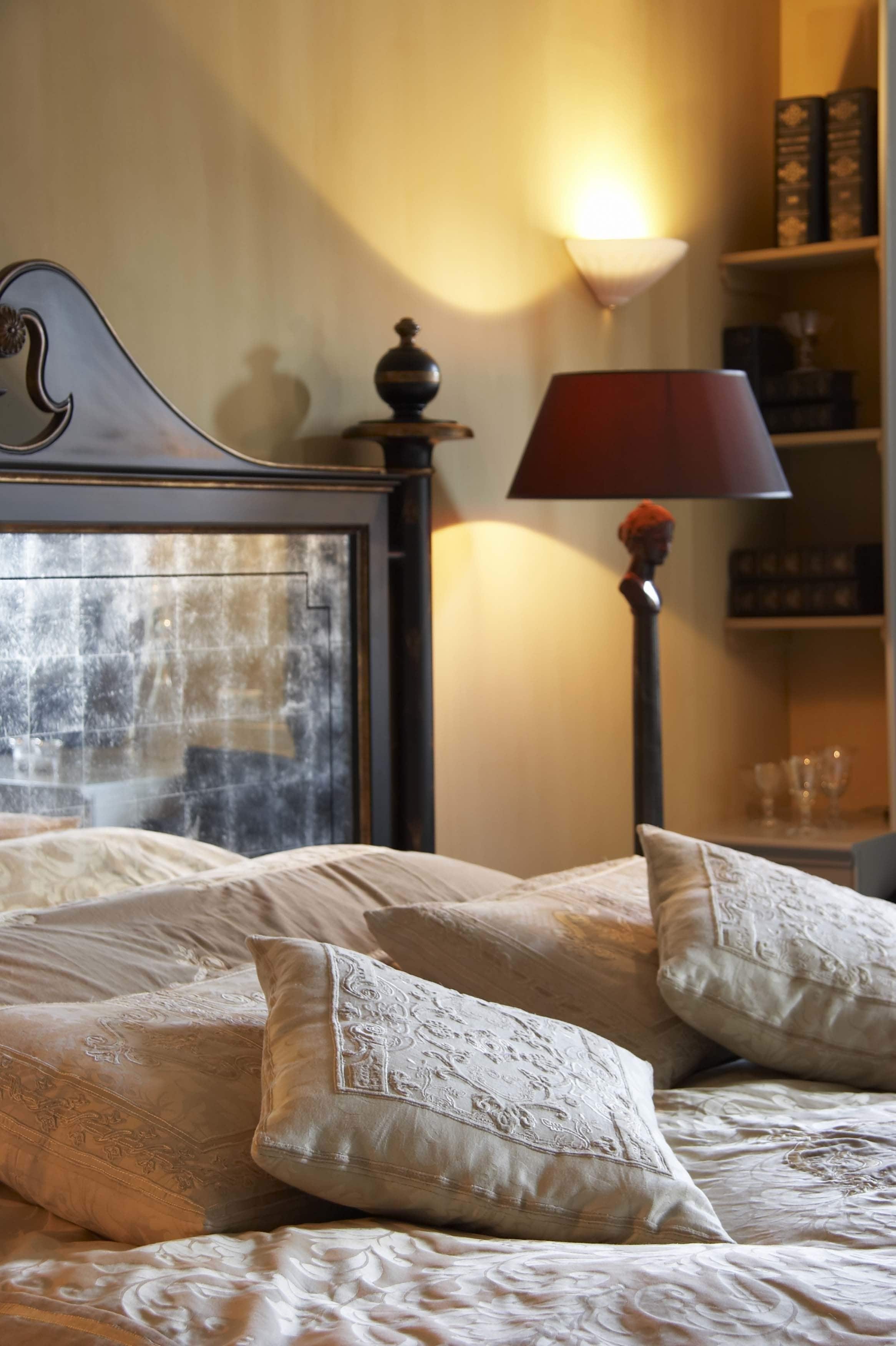 Una cama bien hecha con almohadas ornamentales y un cabecero decorativo, una lámpara de noche y un estante con libros y gafas.