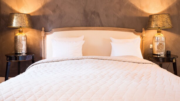 A bed with a white quilted bedspread, flanked by two nightstands with lamps and bottles of Saint-Emil liquor. - Suitehotel Posthoorn (Monnickendam)