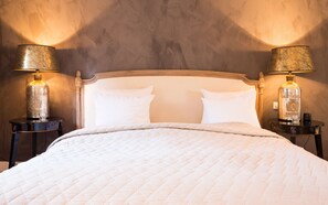 Un lit recouvert d’une courtepointe blanche, flanqué de deux tables de chevet sur lesquelles reposent des lampes et des bouteilles de liqueur de Saint-Émilion.