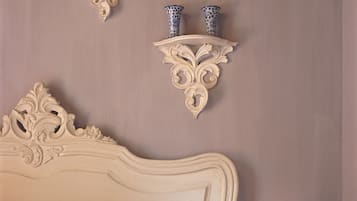 Un dormitorio con una cama blanca clásica, un cabecero decorativo, un cojín estampado en azul y blanco, y una estantería de pared con dos jarrones.