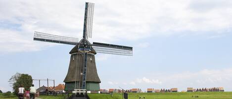 Un molino de viento tradicional con una escalera que lleva a la parte superior, rodeado por un campo cubierto de hierba y una hilera de casas al fondo.