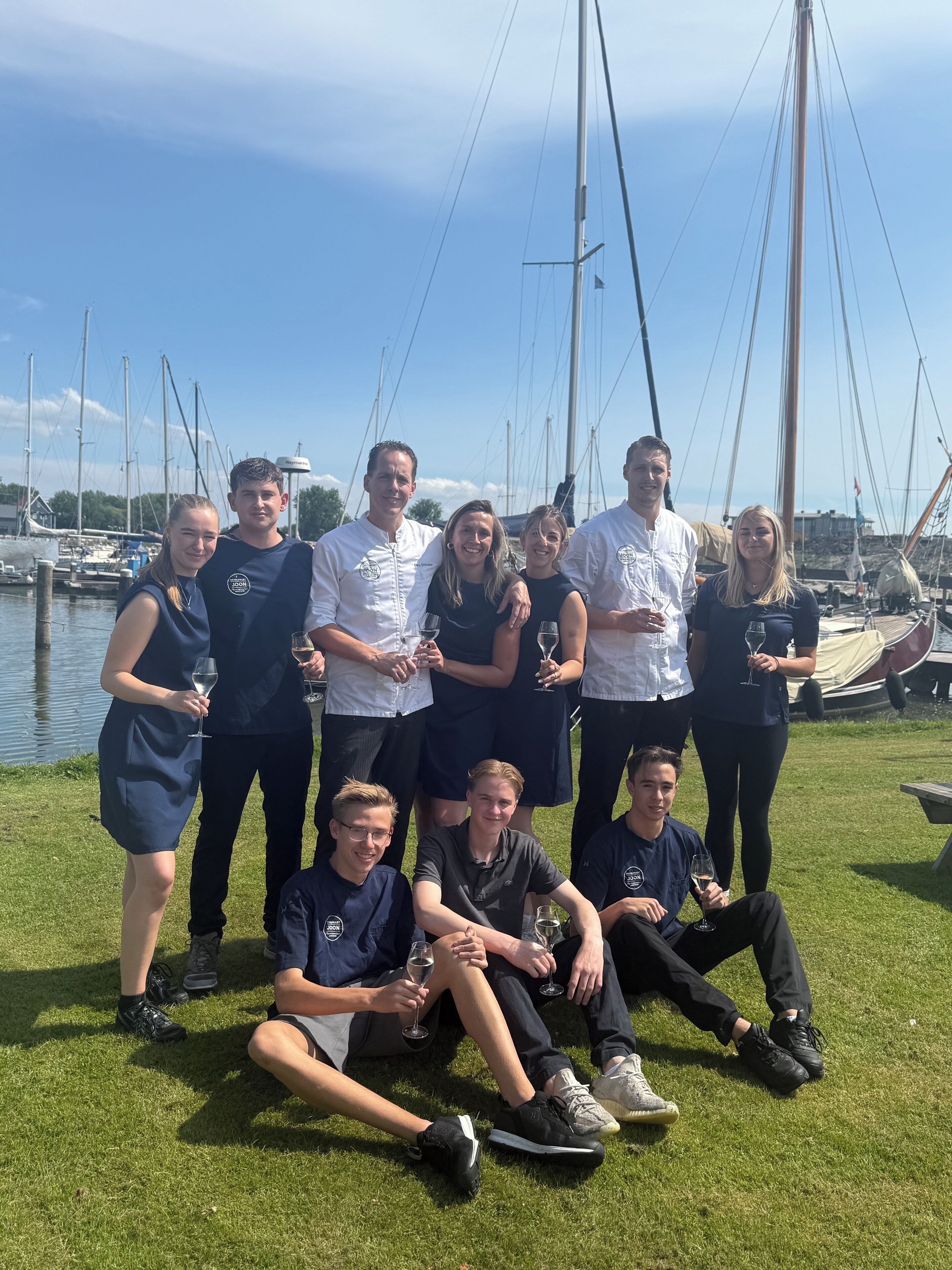 Een groep mensen poseert op een grasveld, met een jachthaven op de achtergrond.