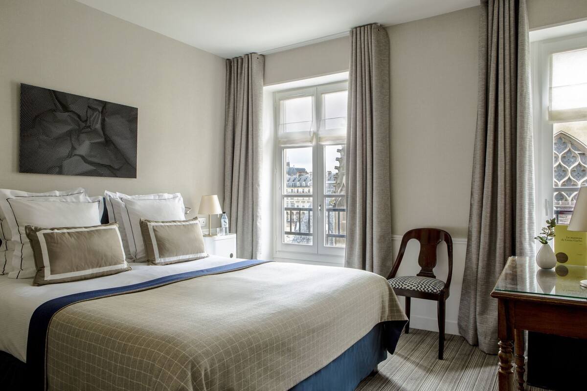 Habitación de hotel con cama, una silla, mesita de noche, ventana con cortinas y un cuadro en la pared.