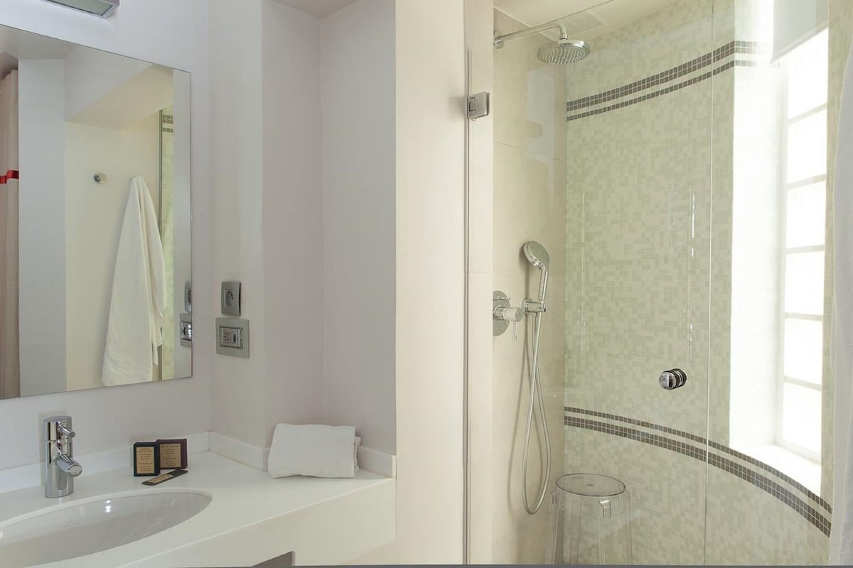 Un baño con un espejo grande, un lavamanos, una ducha con puerta de vidrio y una ventana pequeña.