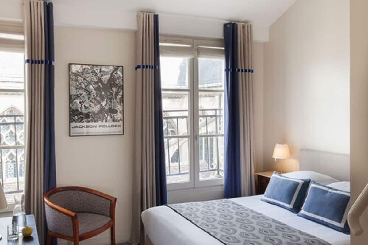 Una habitación de hotel con cama, una silla, una mesa con lámpara y una ventana con cortinas.