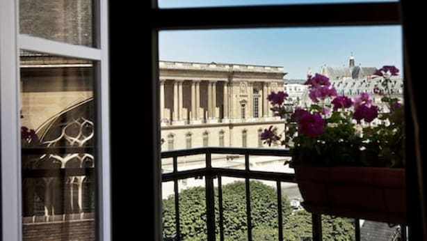 Vista desde una ventana con balcón, mostrando un edificio de arquitectura clásica y una maceta con flores moradas.