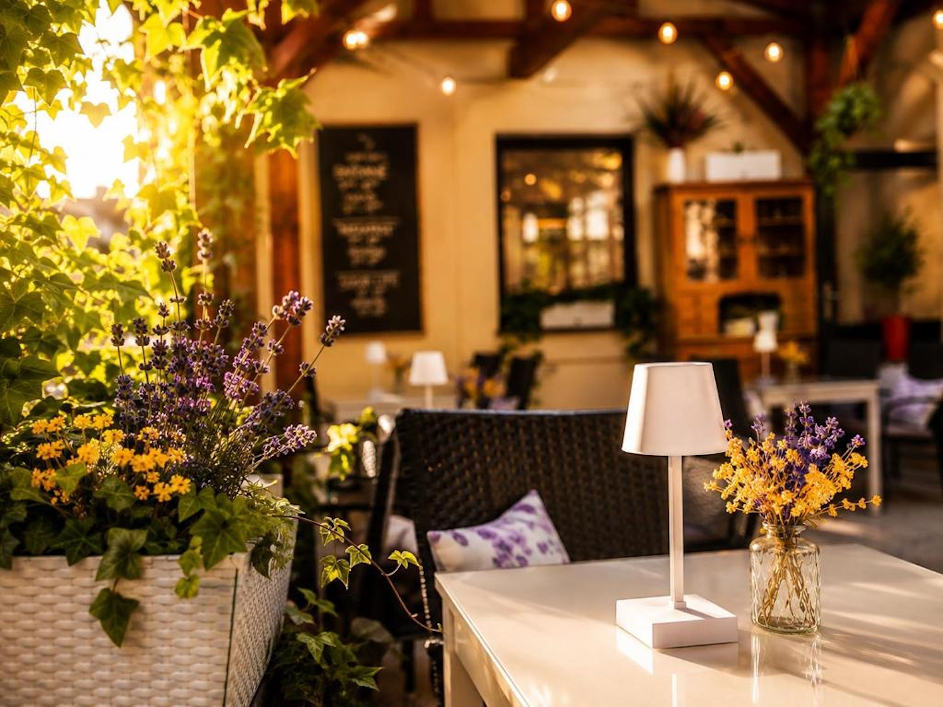 A table with a lamp and a vase of flowers, surrounded by greenery and a cozy interior.