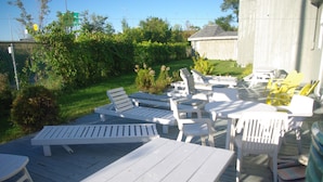 An outdoor deck with white lounge chairs and tables, surrounded by greenery and a wooden fence.