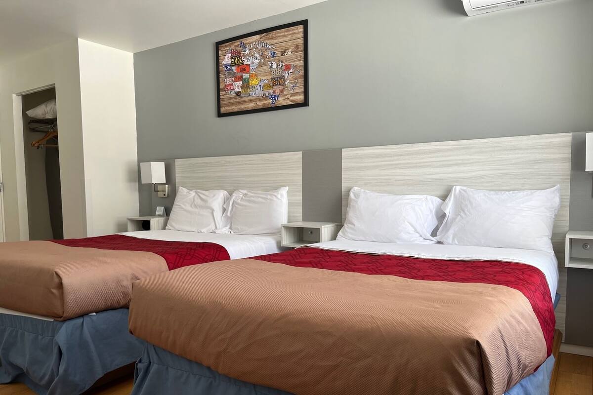 Une chambre d’hôtel avec deux lits, chacun recouvert d’une courtepointe rouge et bleue, avec du linge de lit blanc et une tête de lit en bois. Un tableau est accroché au mur au-dessus des lits, et une petite table de chevet avec une lampe se trouve à côté de chaque lit.