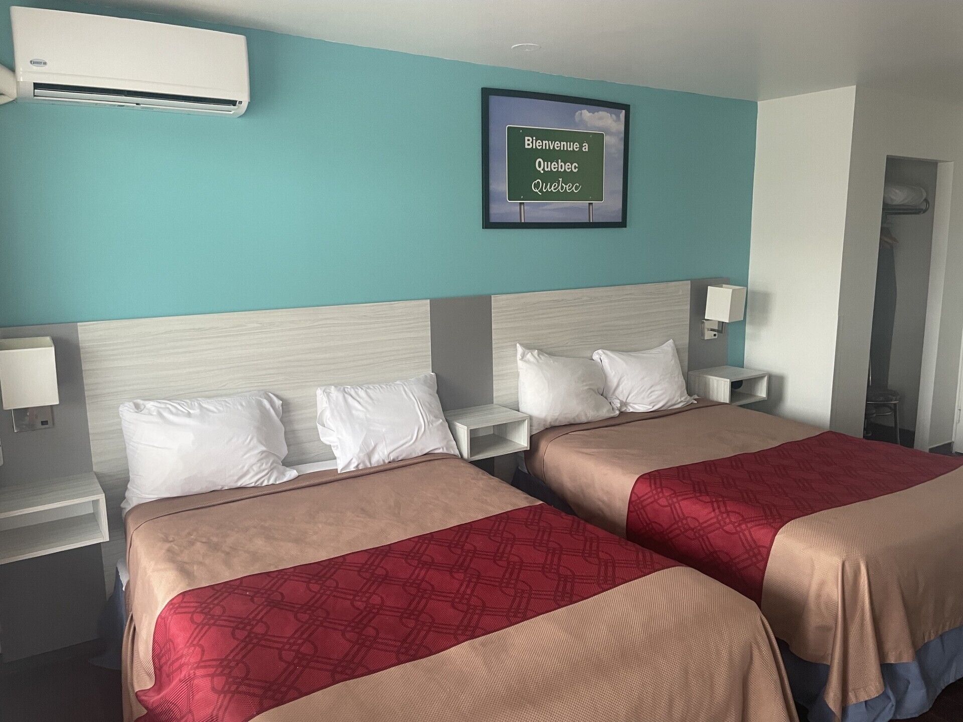Habitación de hotel con dos camas, aire acondicionado de pared, un cartel de bienvenida enmarcado, mesitas de noche con lámparas y un armario.