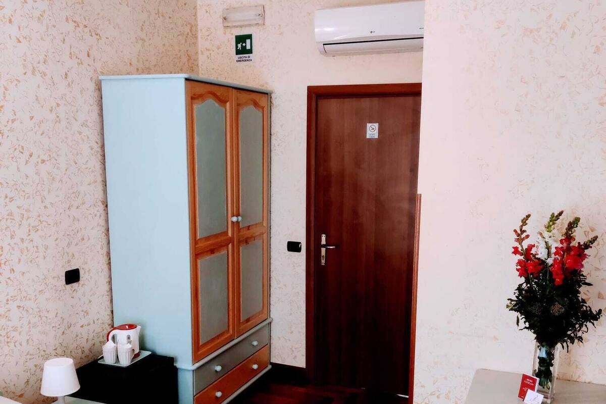 A hotel room with a bed, a wardrobe, a desk with a vase of flowers, and a door with a sign.