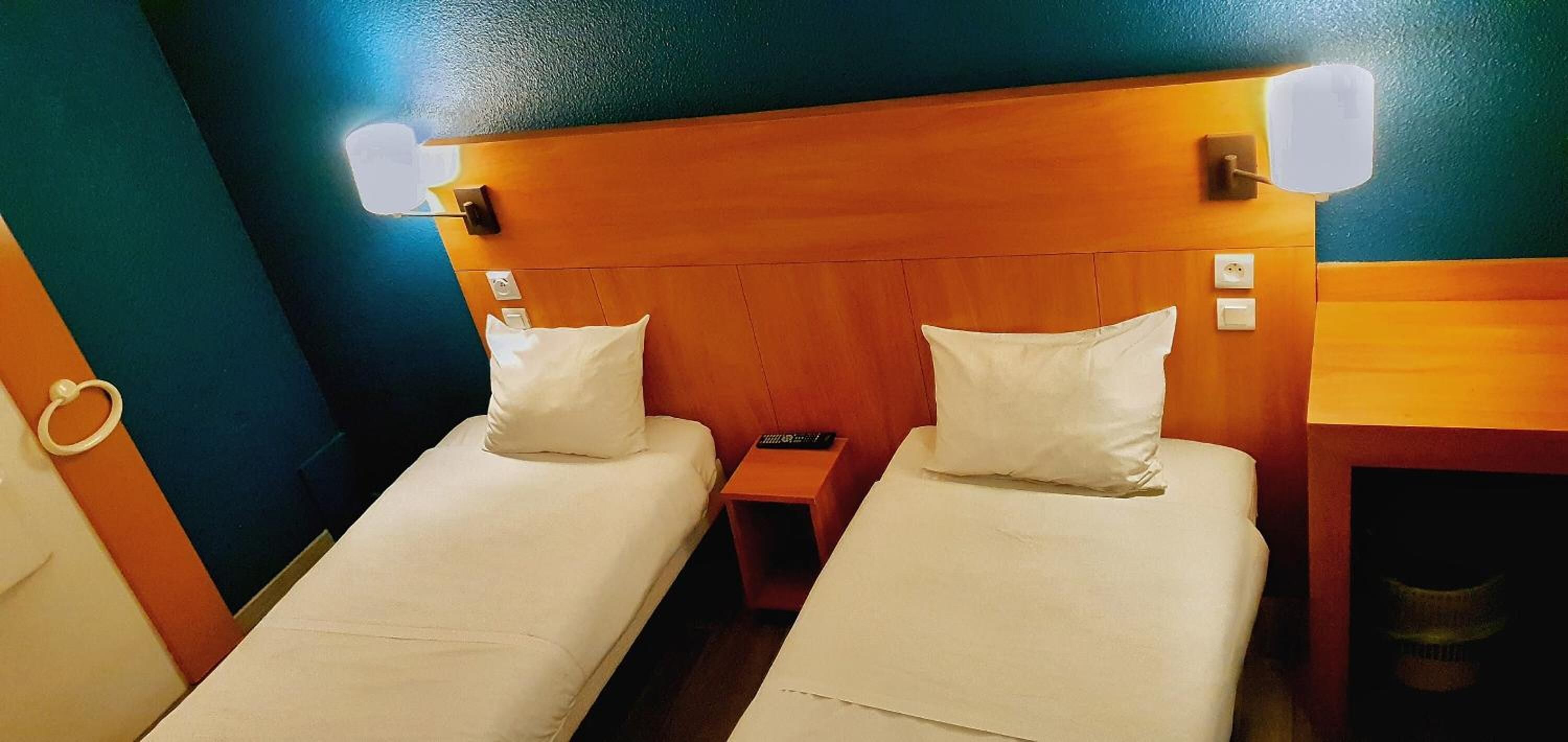 A hotel room with two single beds, wooden headboards, bedside lamps, and a small nightstand.