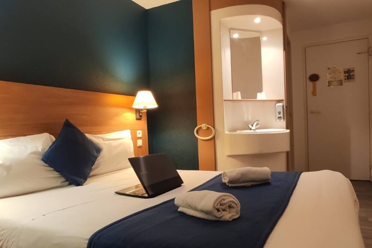 A hotel room with a bed, a laptop, a towel, a nightstand, and a bathroom mirror.