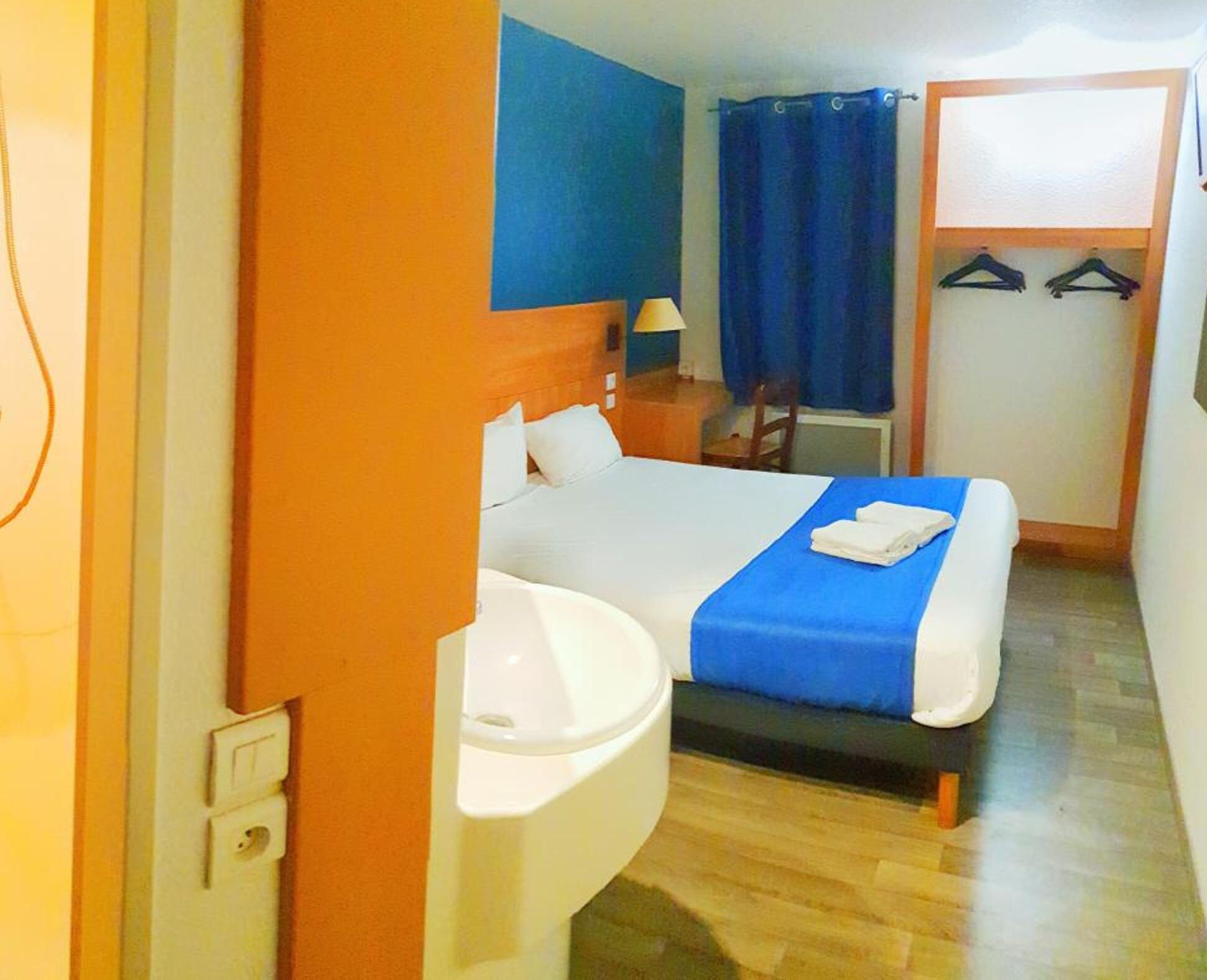 A hotel room with a bed, a sink, a mirror, a chair, a desk, a lamp, a blue curtain, and a coat rack with hangers.