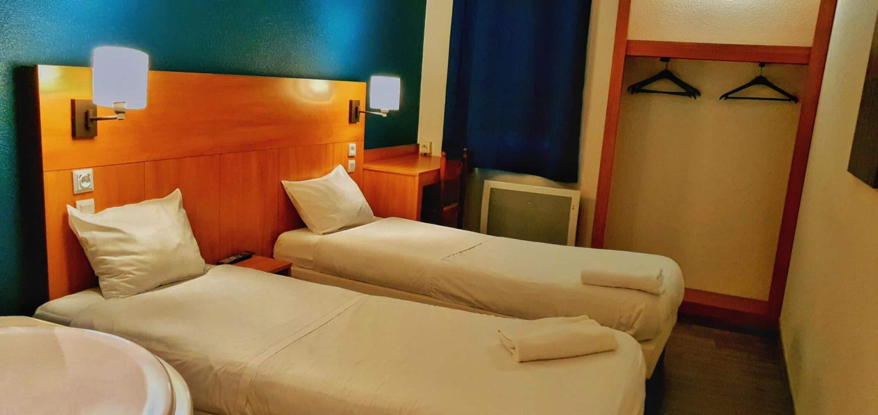 Habitación de hotel con dos camas, cabecero de madera, lámparas de noche, una mesita y un armario con perchas.
