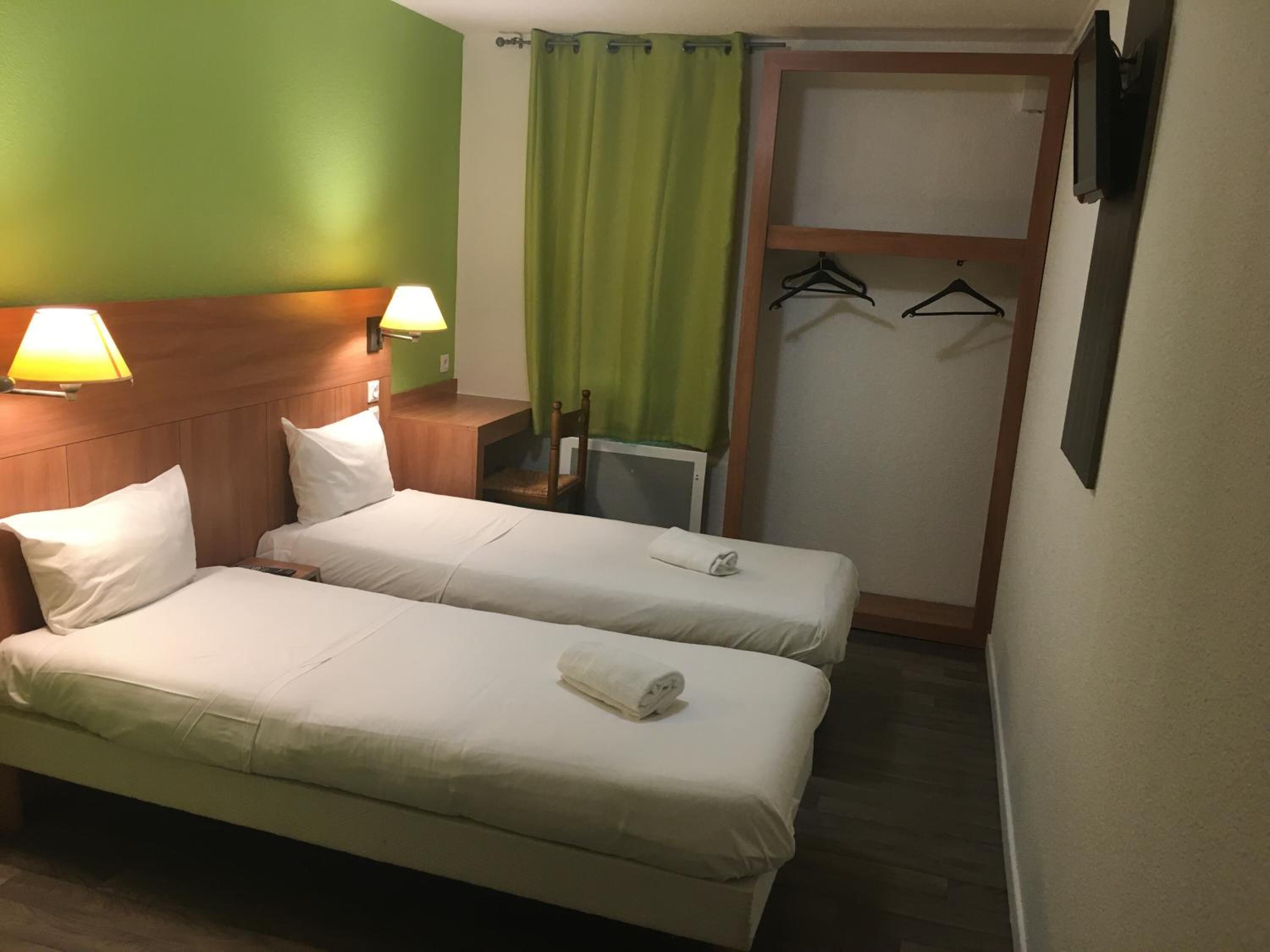 Habitación de hotel con dos camas individuales, cabecera de madera, pared verde, cortina verde y perchero con perchas.