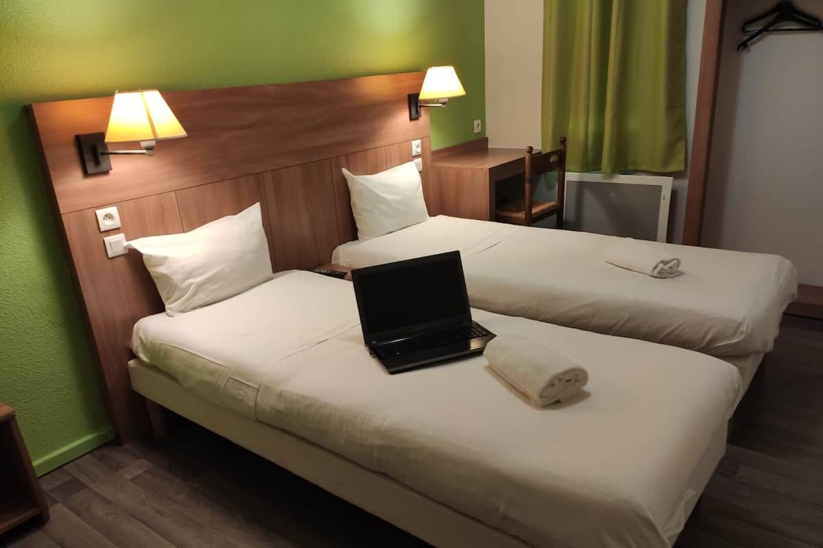 A hotel room with two beds, each with a pillow, a laptop on one bed, and a rolled towel on the other. There are wall-mounted lamps and a small table between the beds.